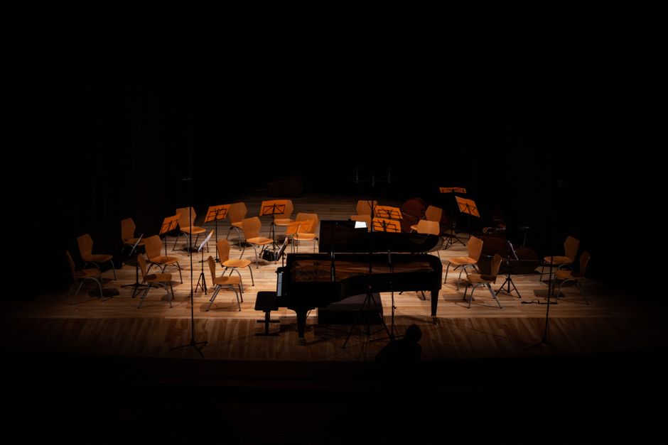 A dimly lit stage set up for a classical music concert featuring empty wooden chairs positioned in front of music stands with sheet music. The chairs are arranged in a semi-circle around a black grand