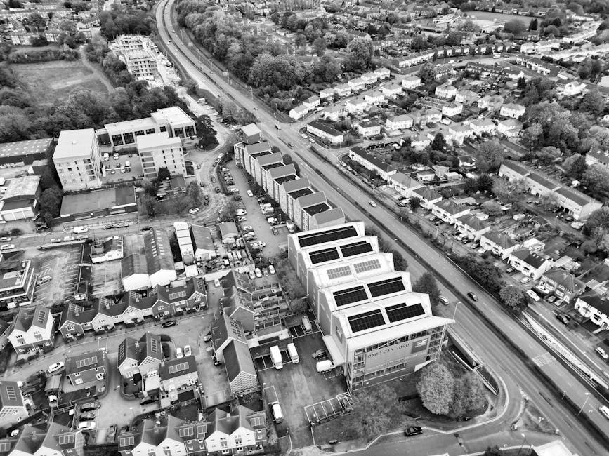 An aerial black-and-white photograph showing a cluster of modern, flat-roofed commercial buildings equipped with solar panels, located alongside a busy urban street with parked vehicles in the loading